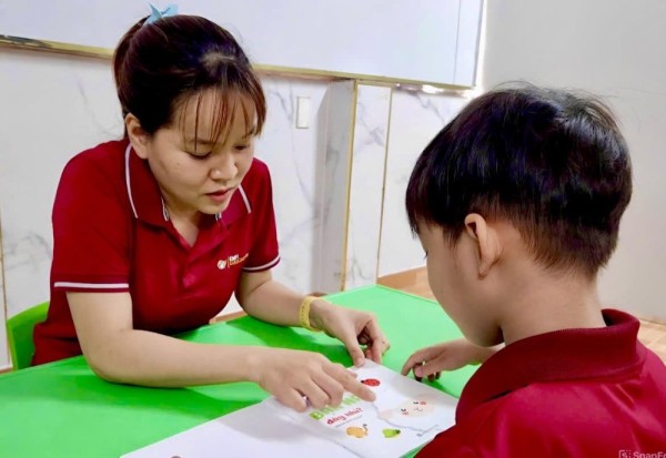 Giáo viên đang tương tác ngôn ngữ với trẻ bằng sách tranh hoặc đồ chơi giáo dục.